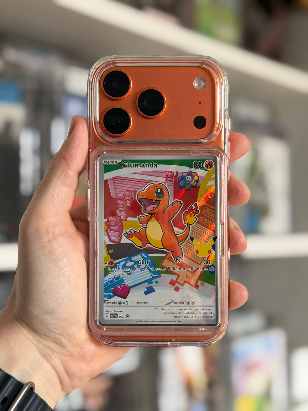 A hand holding a clear phone case with a built-in card slot displaying a Charmander Pokémon card, set against a blurred indoor background.